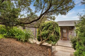 A modern house with a wooden deck, surrounded by lush greenery and a large tree.