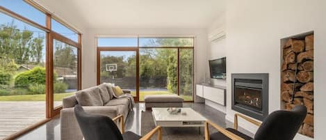 A modern living room with a sofa, chairs, a coffee table, a fireplace, and large windows.
