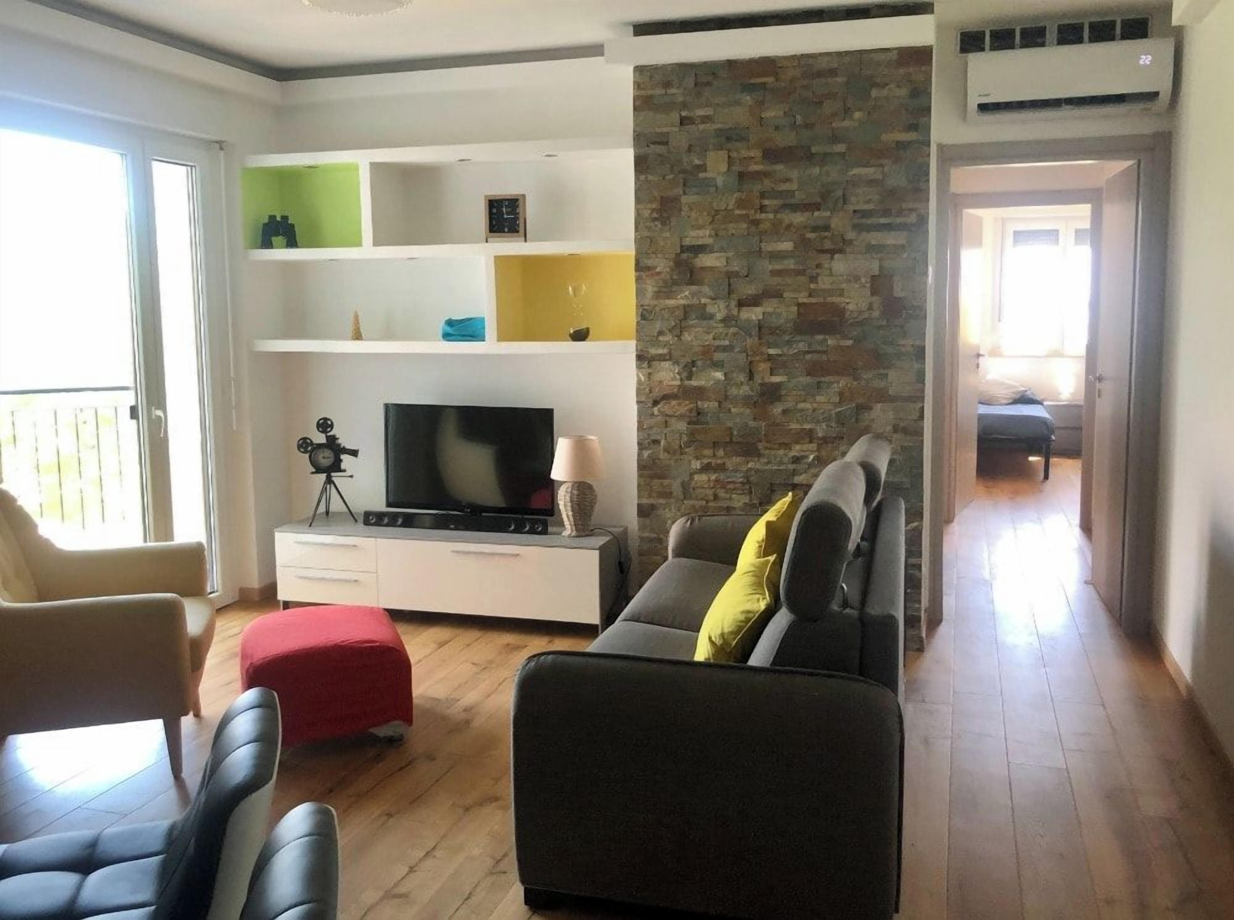 Un salon avec un canapé, un pouf, une télévision et des étagères.