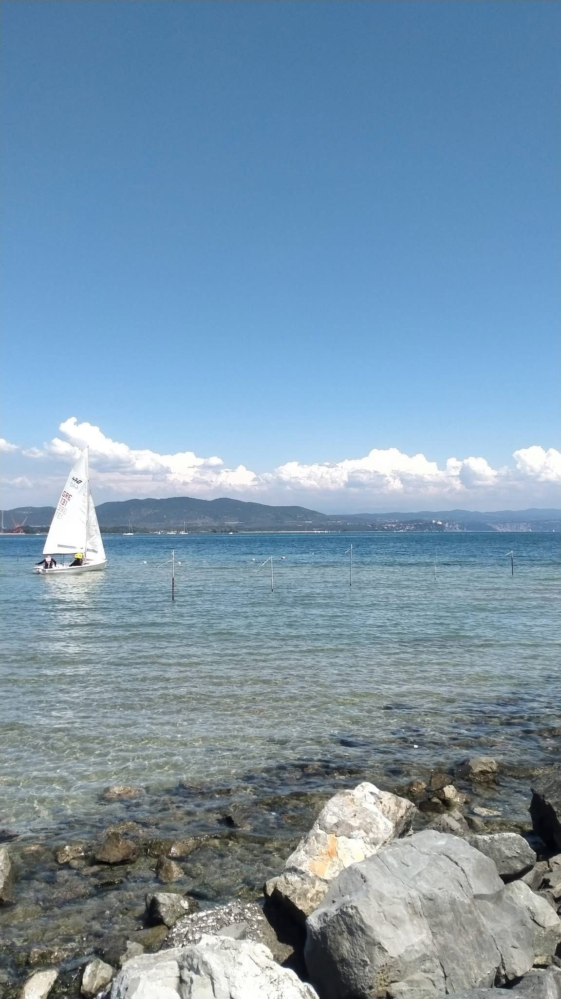 Un voilier sur une étendue d’eau calme, sous un ciel dégagé, avec des montagnes qui se dessinent à l’horizon.