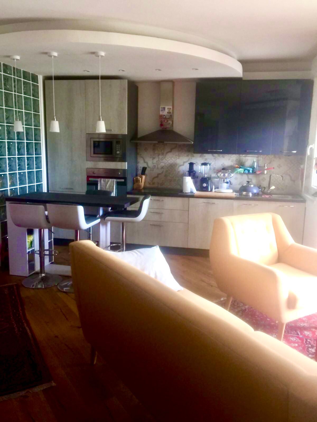 A modern kitchen with a microwave, stove, and sink. There are two bar stools and a dining table with chairs. The room has a wooden floor and a large window.