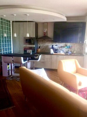 A modern kitchen with a microwave, stove, and sink. There are two bar stools and a dining table with chairs. The room has a wooden floor and a large window.