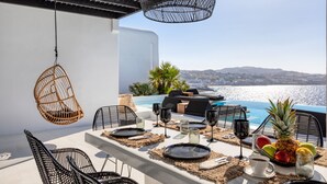 An outdoor dining area with a view of the sea, featuring a table set with plates, glasses, and fruits. - Psarou Black Villa II (Mykonos)