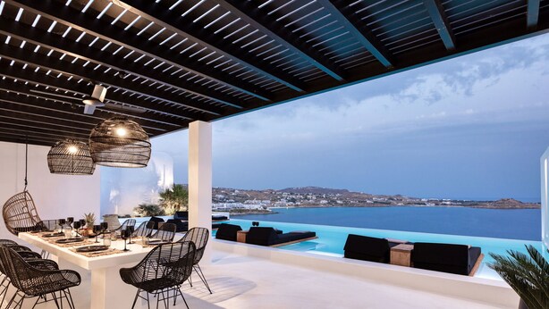 An outdoor dining area with a view of the sea, featuring a white table, black chairs, and hanging lights. - Psarou Black Villa II (Mykonos)