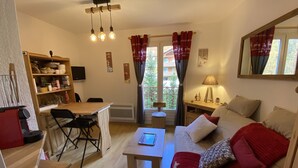 A cozy living room with a wooden dining table, a sofa with red and white cushions, a coffee table, and a TV mounted on the wall. - STUDIO CENTER RESORT LA FOUX D'ALLOS SLEEPS 4 MOUNTAIN CHALET ATMOSPHERE (La Foux d’Allos)
