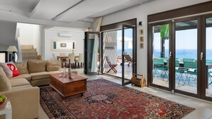 A modern living room with a beige sofa, a wooden coffee table, and a patterned rug. There is a sliding glass door leading to an outdoor area with a view of the sea. - Villa Athena Lindos - 6 Bedroom Villa with Private Infinity Pool and Seaviews (Lindos)