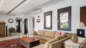 A living room with a beige sofa, a wooden coffee table, and a red patterned rug. There is a large window with a view of a mountain. - Villa Athena Lindos - 6 Bedroom Villa with Private Infinity Pool and Seaviews (Lindos)