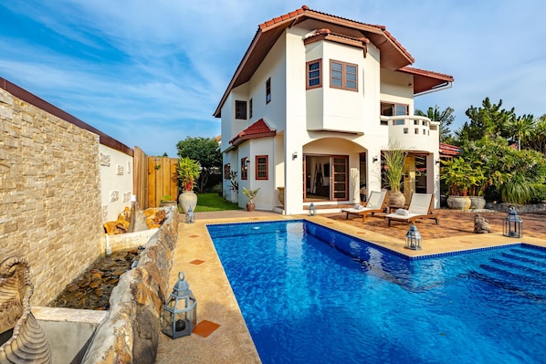 Una casa de dos pisos con piscina, paredes de piedra y mobiliario para exteriores.
