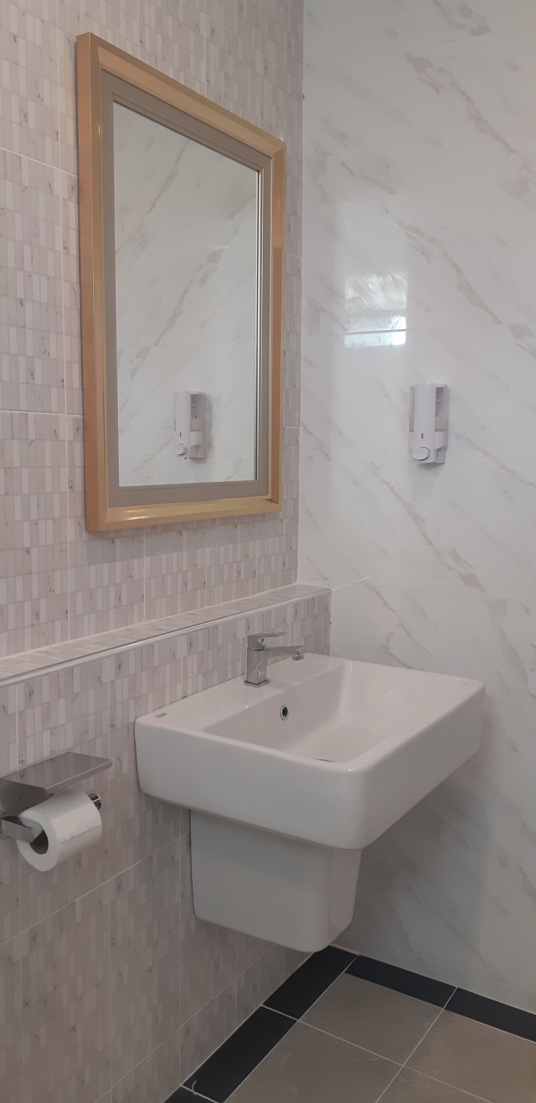 Un baño con pared de mármol, un lavamanos, un espejo con marco de madera y un dispensador de papel higiénico.