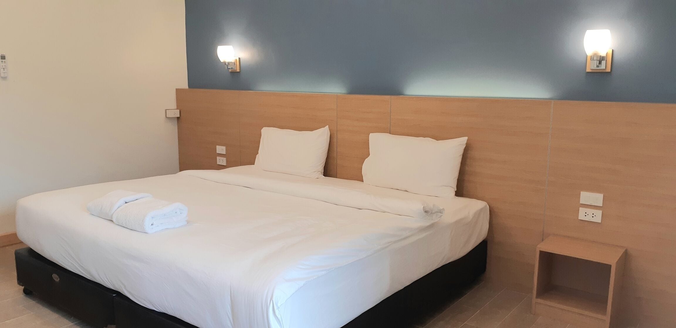 Cama bem arrumada com lençóis brancos e toalha dobrada, cabeceira de madeira e duas luminárias fixadas na parede.