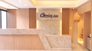 Zona de recepción de un hotel con un mostrador de mármol, paneles de madera y un cartel que dice 'Cenil Hotel Lampang'.