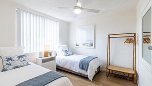 Une chambre avec deux lits, un ventilateur de plafond, une table de chevet avec une lampe, un tableau encadré au mur et un étendoir à linge en bois.