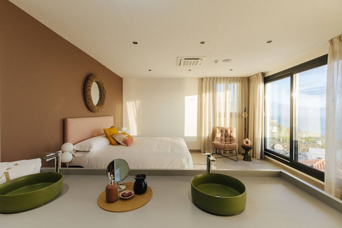 Une chambre d’hôtel moderne dotée d’un grand lit, d’un coin salon avec un fauteuil, d’une vue sur la ville et d’une salle de bain avec deux lavabos.