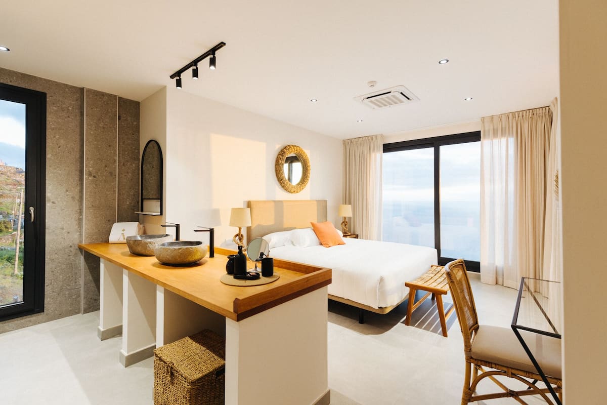 Une chambre d’hôtel moderne avec un grand lit, un bureau en bois, un fauteuil en osier et une vue sur la ville.