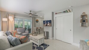 Un salon moderne comprenant un canapé, un fauteuil, une table basse et un coin repas avec une table et des chaises. On y trouve également un ventilateur de plafond, un tableau représentant des pélicans et une horloge au mur.