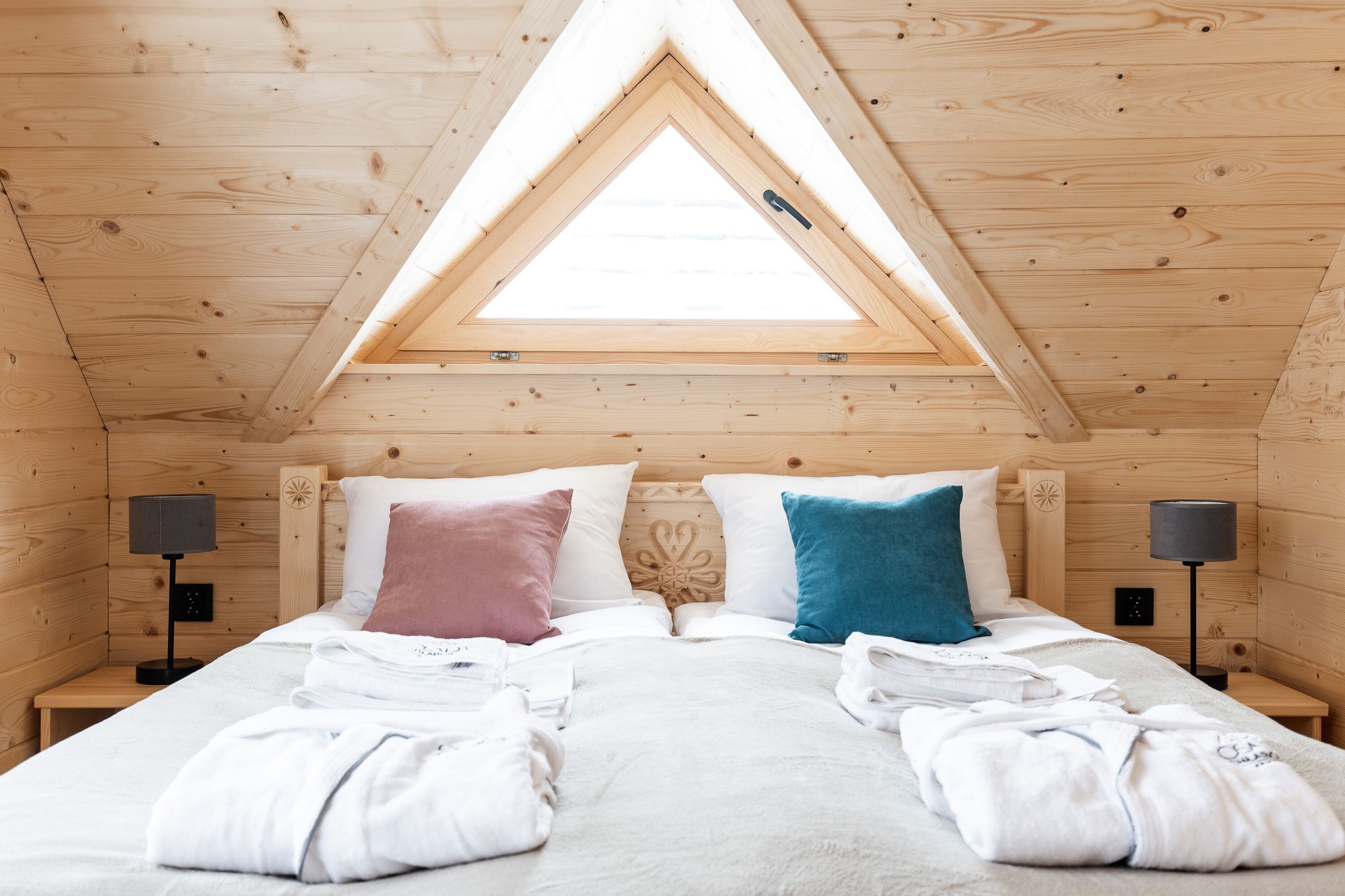 Quarto de cabana de madeira com claraboia, duas mesas de cabeceira e uma cama com almofadas.