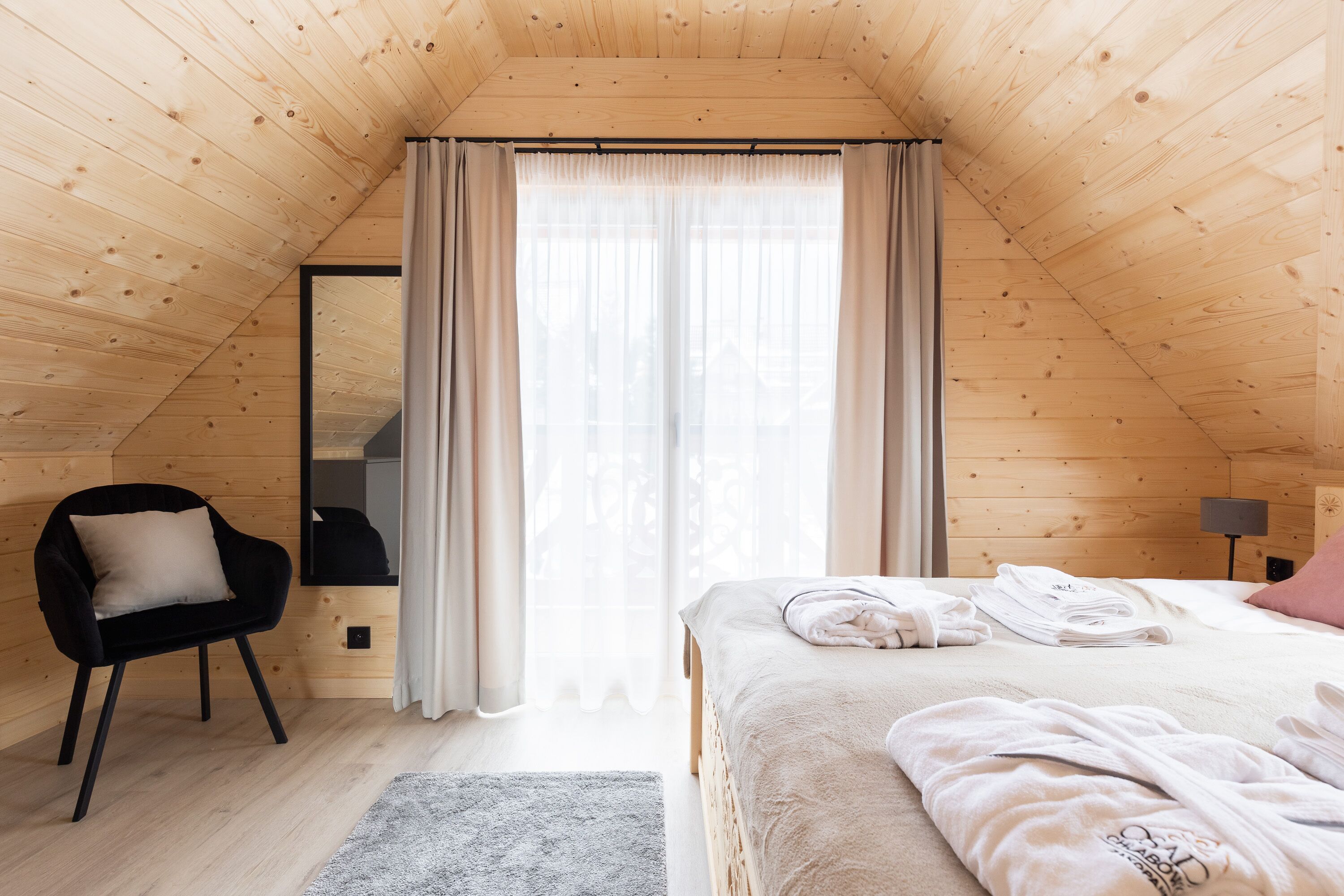 A cozy room with a sloped ceiling, a bed with neatly folded towels, a black chair with a cushion, and a small table with a lamp.
