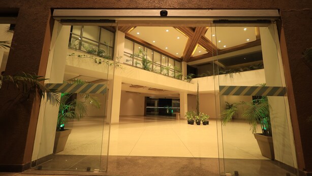 A spacious lobby with a high ceiling, glass doors, and multiple potted plants. - Heaven7 Hotel & Resort (Himatnagar)