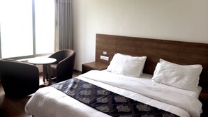 A hotel room with a large bed, a bedside table, a chair, and a small table. - Heaven7 Hotel & Resort (Himatnagar)