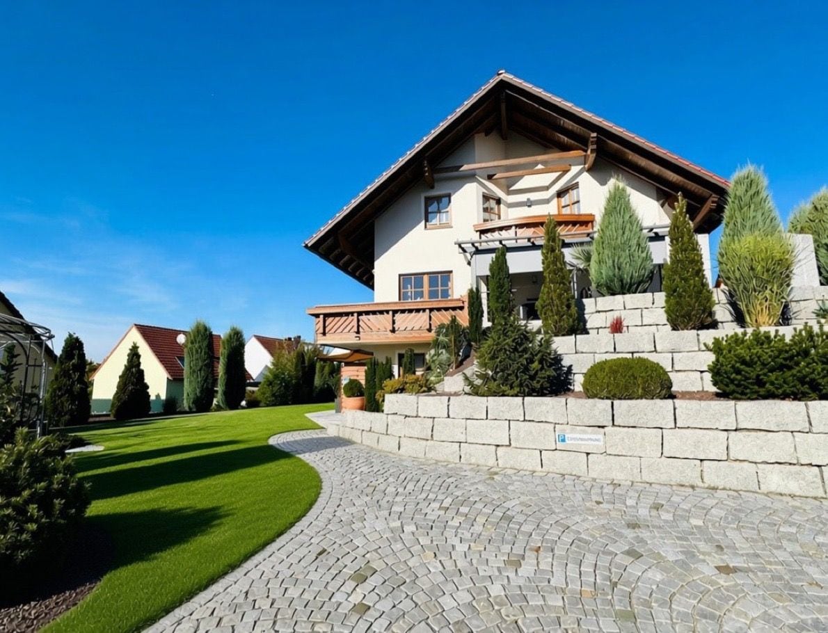 A two-story building with a stone pathway leading to the entrance, surrounded by well-maintained greenery and a clear blue sky.
