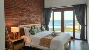 Ein Hotelzimmer mit Backsteinwand, einem Bett mit weißer Bettwäsche und grünen Kissen, einem Holz-Nachttisch, einer Lampe und Blick auf den Strand durch eine Glastür.
