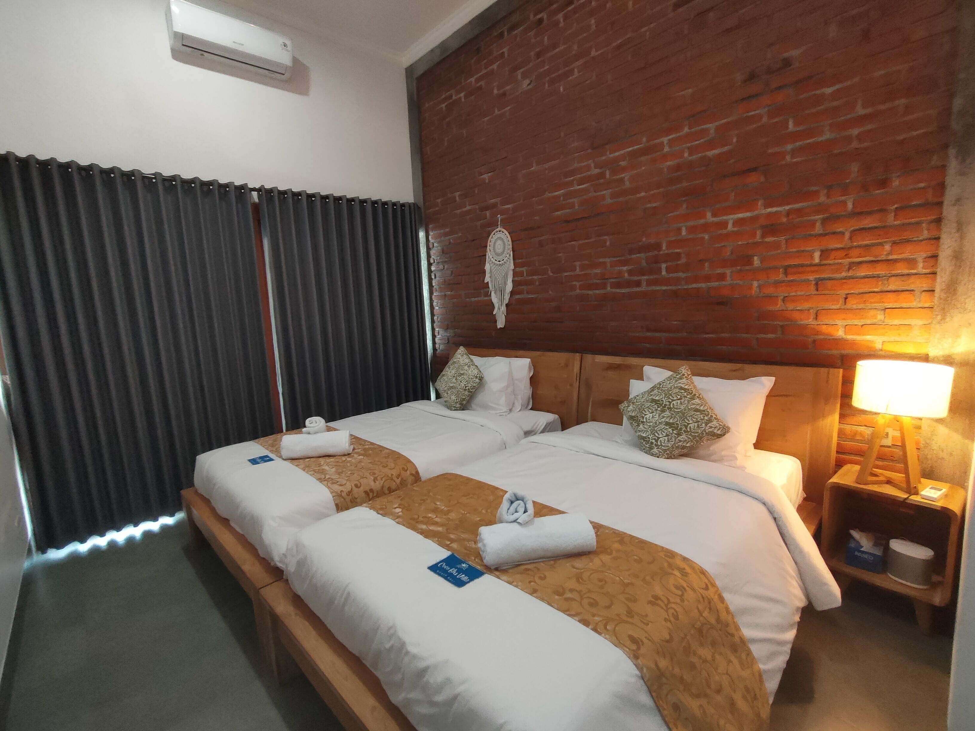 Habitación de hotel con cama doble, cabecera de madera, pared decorativa de ladrillo, mesita de noche con lámpara y ventana con cortinas.
