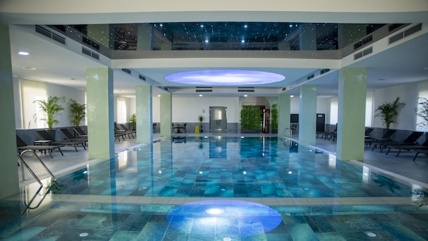 Una gran piscina interior con un estampado de cuadros en el suelo, rodeada de sillones de descanso y plantas en macetas.