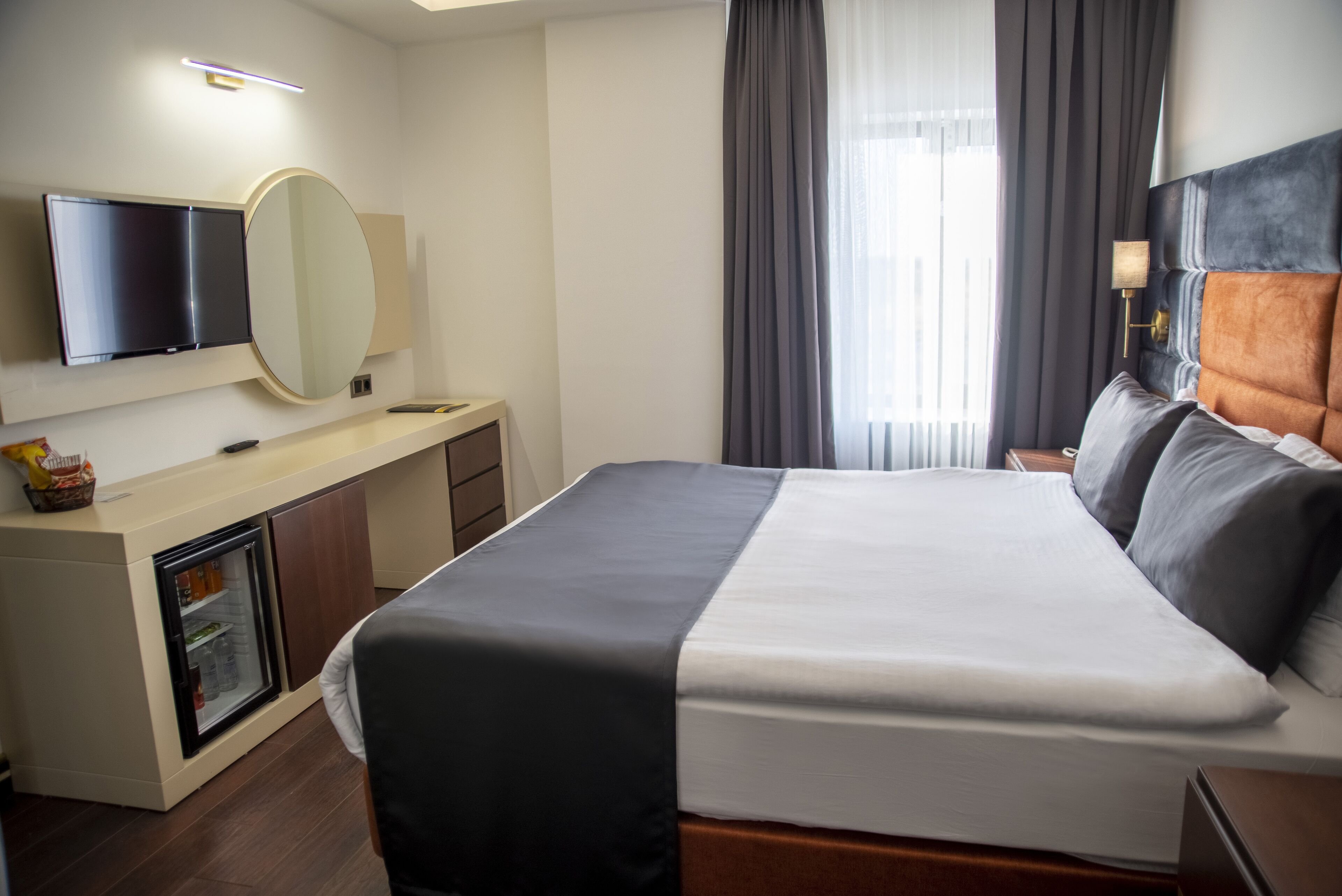 A hotel room with a bed, a TV, a mirror, a small refrigerator, and a window with curtains.