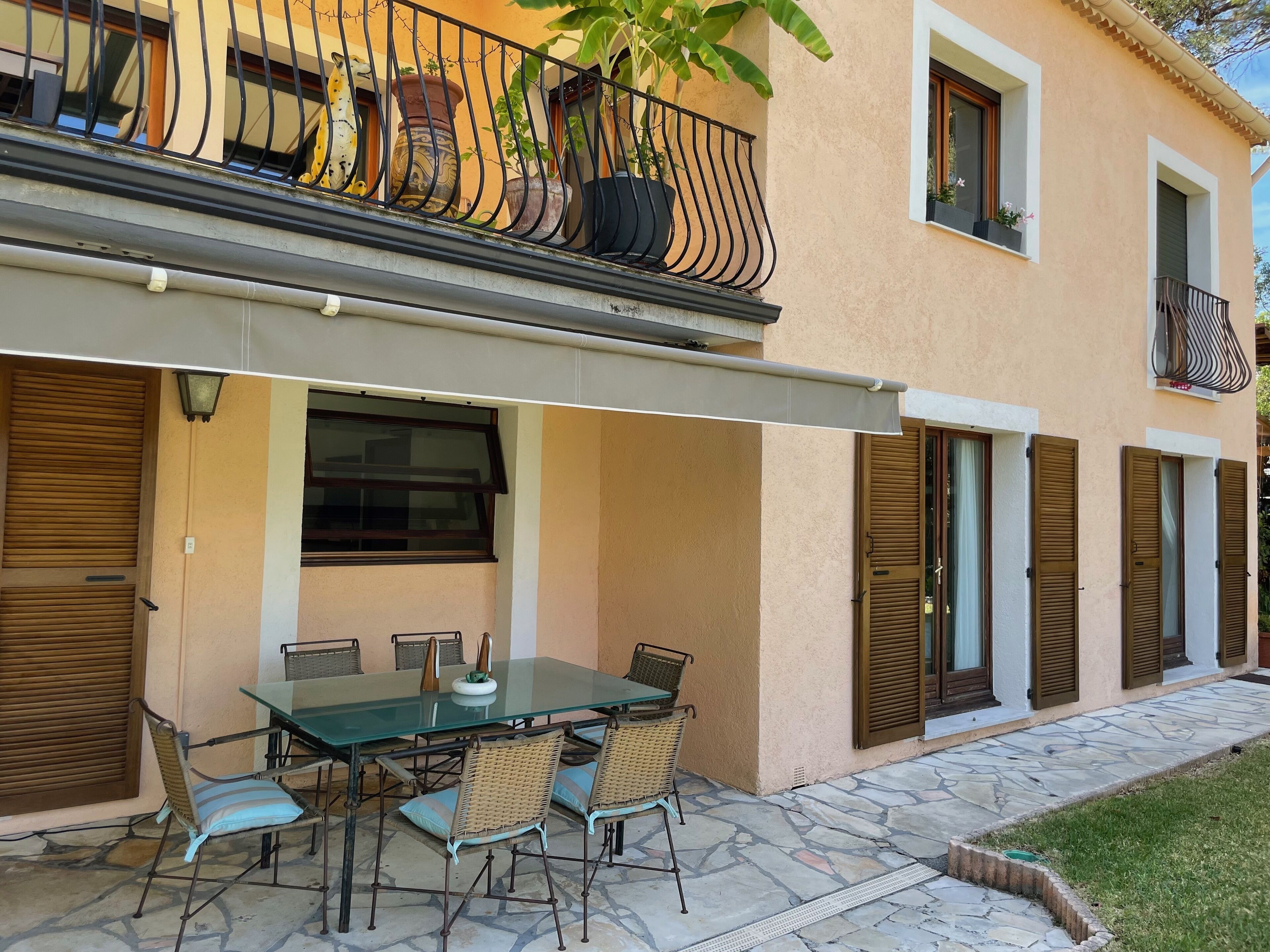 Un espace extérieur aménagé pour les repas, avec une table en verre et des chaises en osier, une terrasse couverte et un balcon agrémenté de plantes en pot.