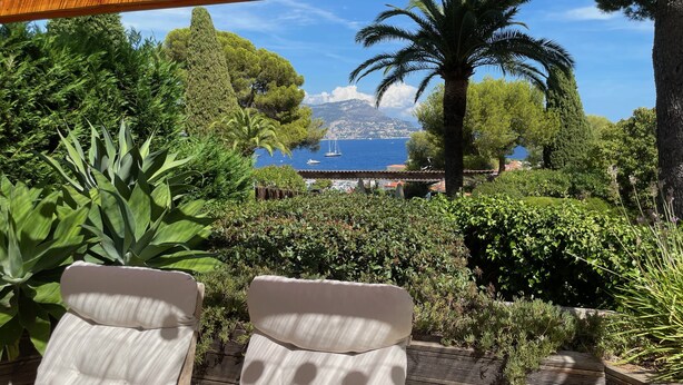 Un patio con due poltrone bianche imbottite, un pergolato in legno e vista sul mare con barche e una montagna all'orizzonte.