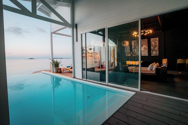 Une piscine à débordement moderne offrant une vue dégagée sur la mer, un coin salon confortable avec un canapé et une table, et un espace intérieur bien éclairé agrémenté d’œuvres d’art aux murs.