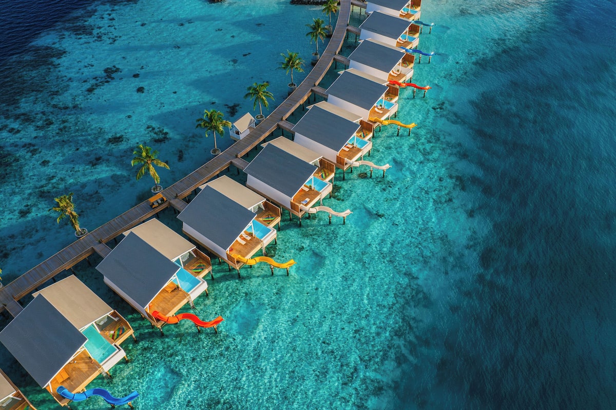 An aerial view of overwater bungalows with clear turquoise water, wooden walkways, and palm trees.