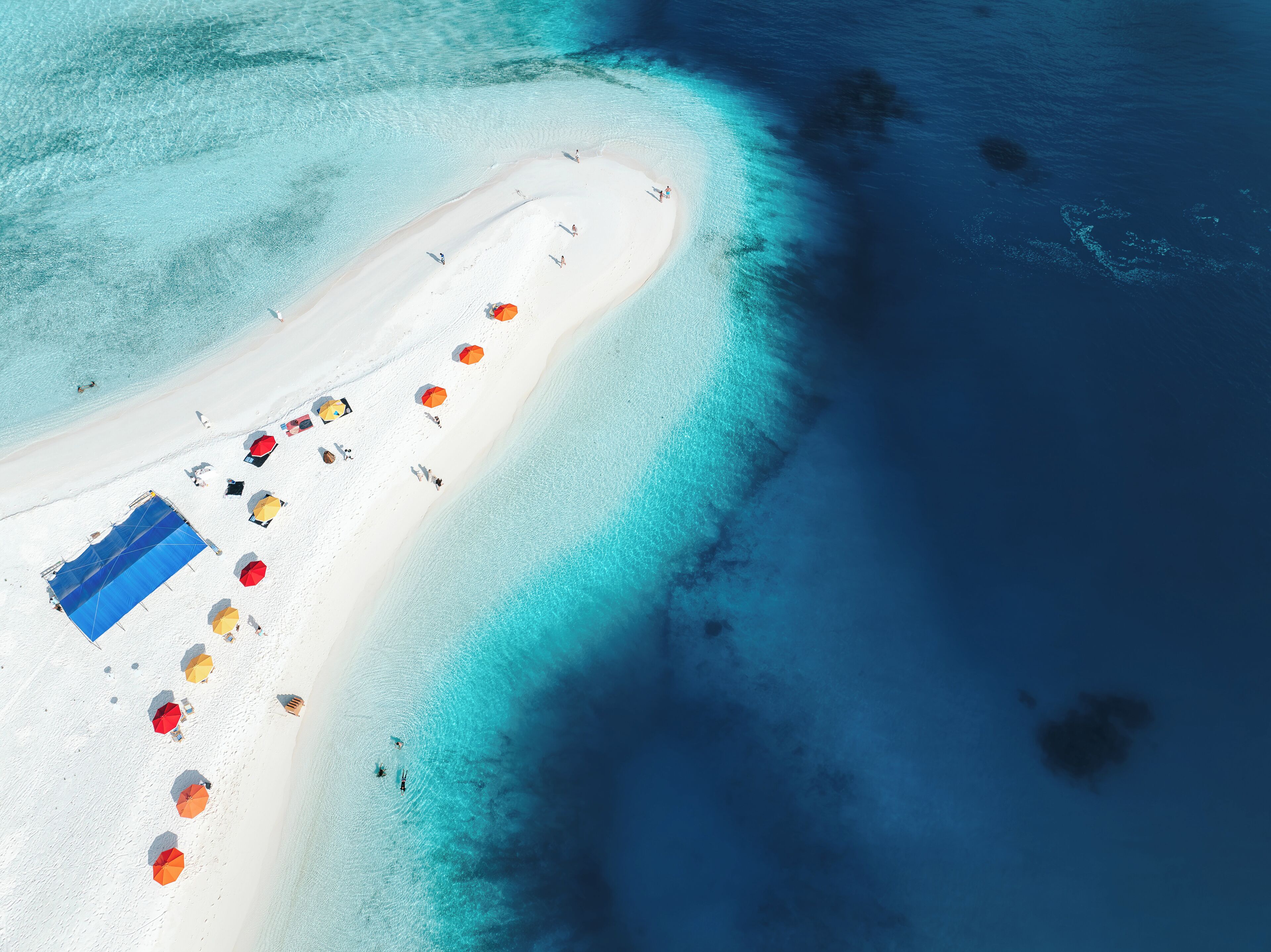 Vista aérea de uma praia com uma extensa faixa de areia branca, águas turquesa cristalinas e diversos guarda-sóis coloridos.