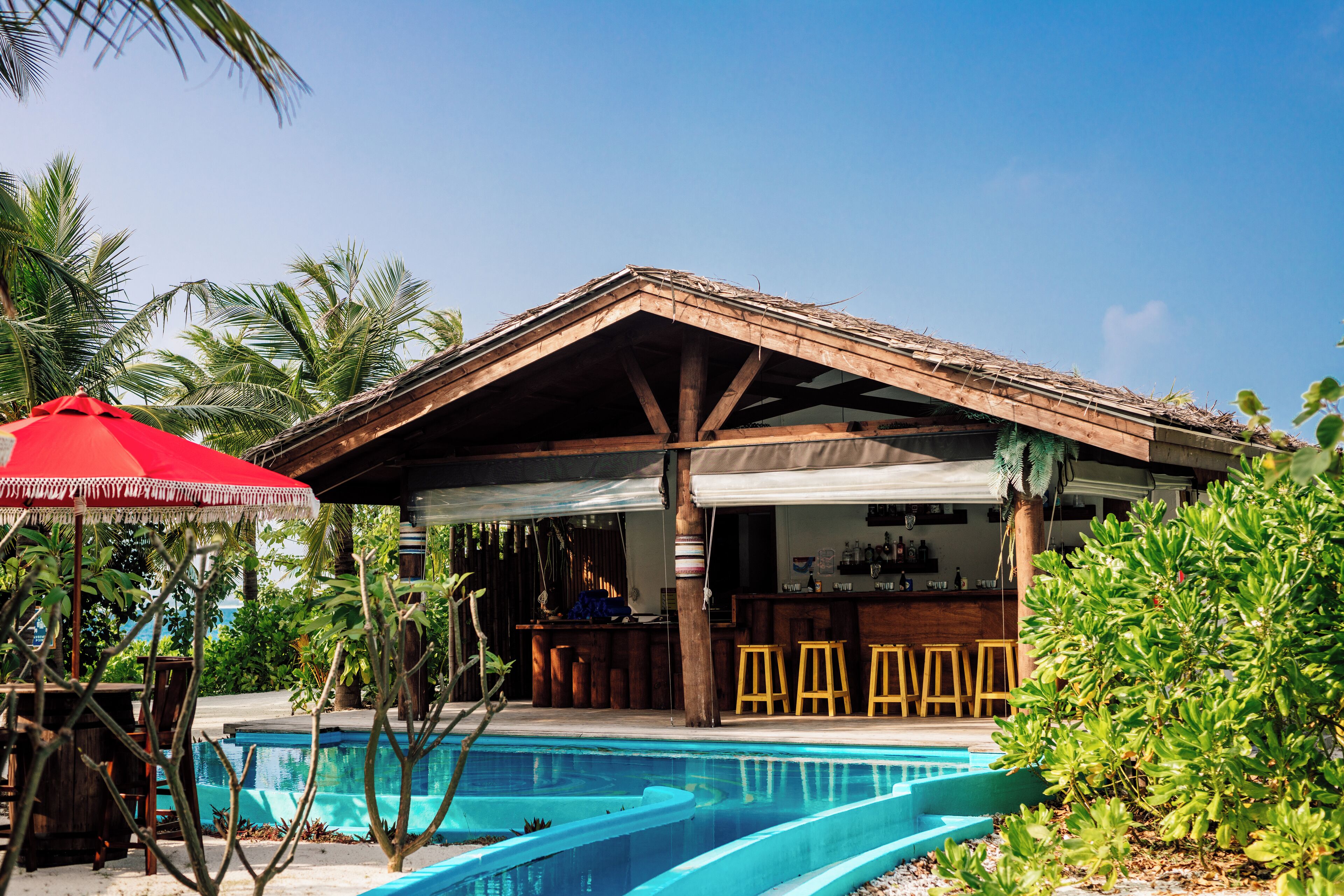 A poolside bar area with wooden stools, a red umbrella, and a pool with clear blue water.