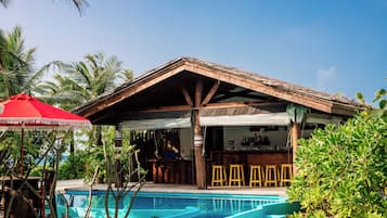 A poolside bar area with wooden stools, a red umbrella, and a pool with clear blue water.