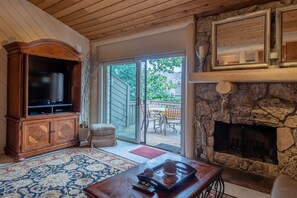 A cozy living room with a stone fireplace, a wooden entertainment center, and a large sliding glass door leading to an outdoor patio. - The Ridge Condo 2617 - Sun Valley Elkhorn 2BR with Fireplace (Sun Valley)