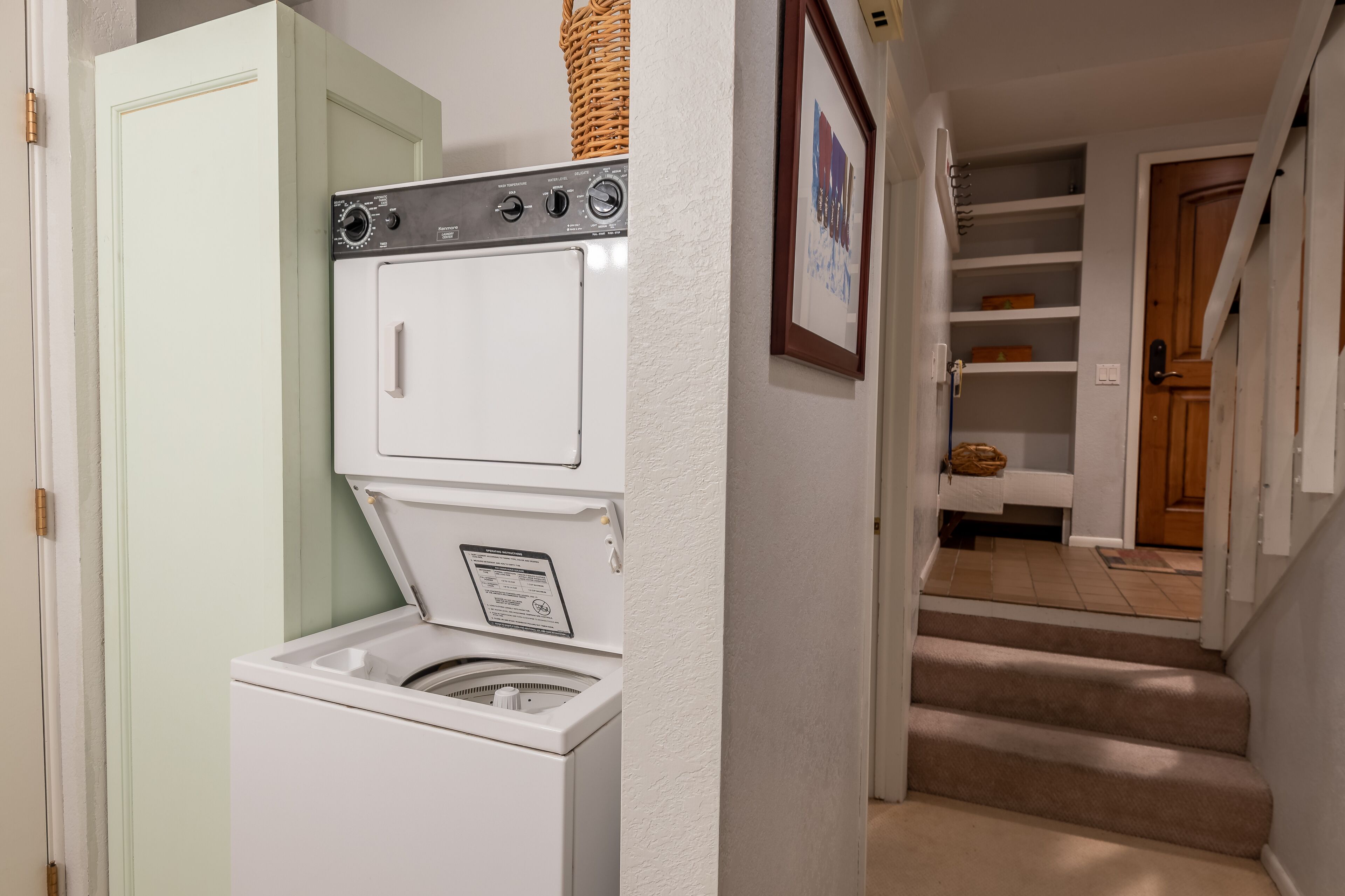 A laundry room with a front-loading washer and dryer, a wicker basket on top of the dryer, and a framed picture on the wall.