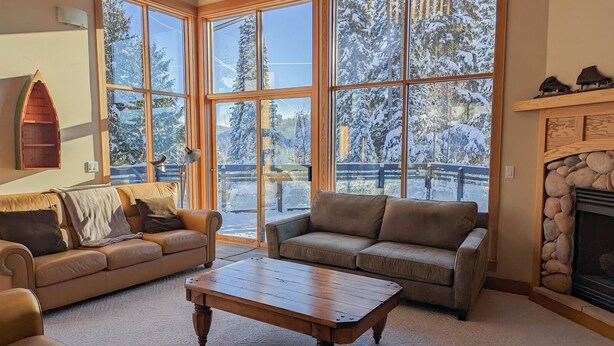 A cozy living room with two sofas and a coffee table, large windows with a view of snow-covered trees, and a stone fireplace.