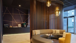 A modern lounge area with a curved sofa, a small round table, and a yellow armchair. A coffee machine and a snack bar are visible in the background. - Dorint Hotel München/Garching (Garching)