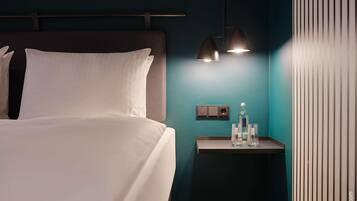 A modern hotel room with a neatly made bed, a nightstand with a water bottle and glasses, and a wall-mounted power outlet.