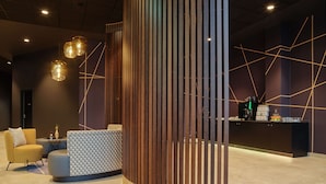 A modern reception area with a curved reception desk, a seating area with a yellow armchair and a patterned ottoman, and a coffee station with a branded coffee machine. - Dorint Hotel München/Garching (Garching)