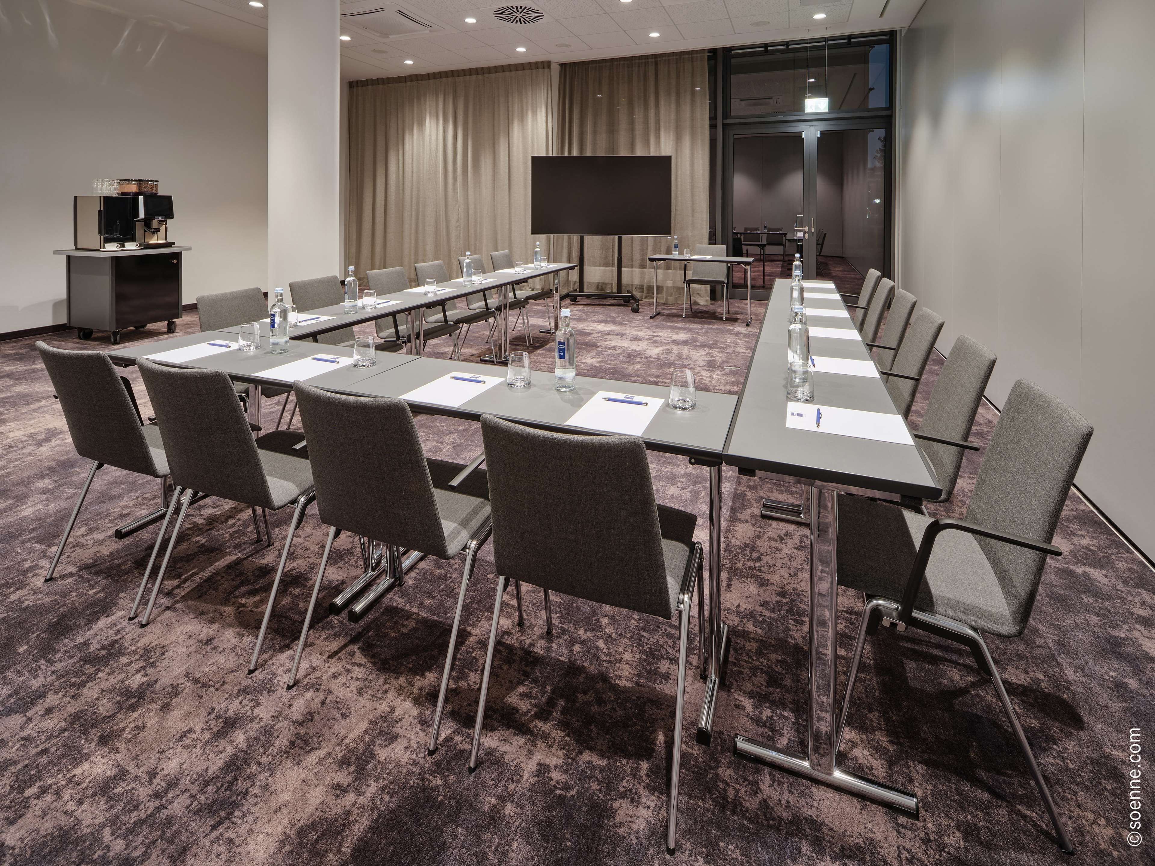 Ein Konferenzraum mit langem Tisch, Stühlen, einer Projektionsleinwand und einer Kaffeemaschine.