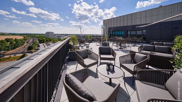 An outdoor terrace with wicker furniture, a view of the city, and a modern building in the background. - Dorint Hotel München/Garching (Garching)