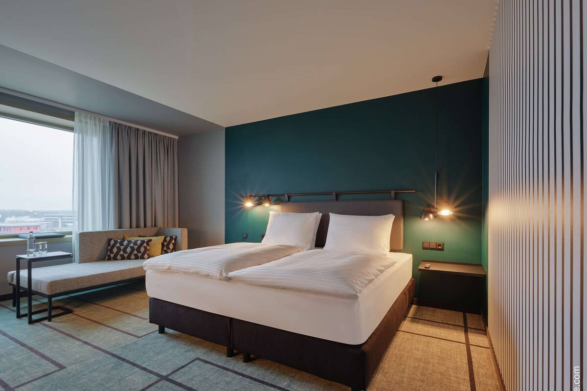 A modern hotel room with a large bed, a sofa, a nightstand, and a view of the city.