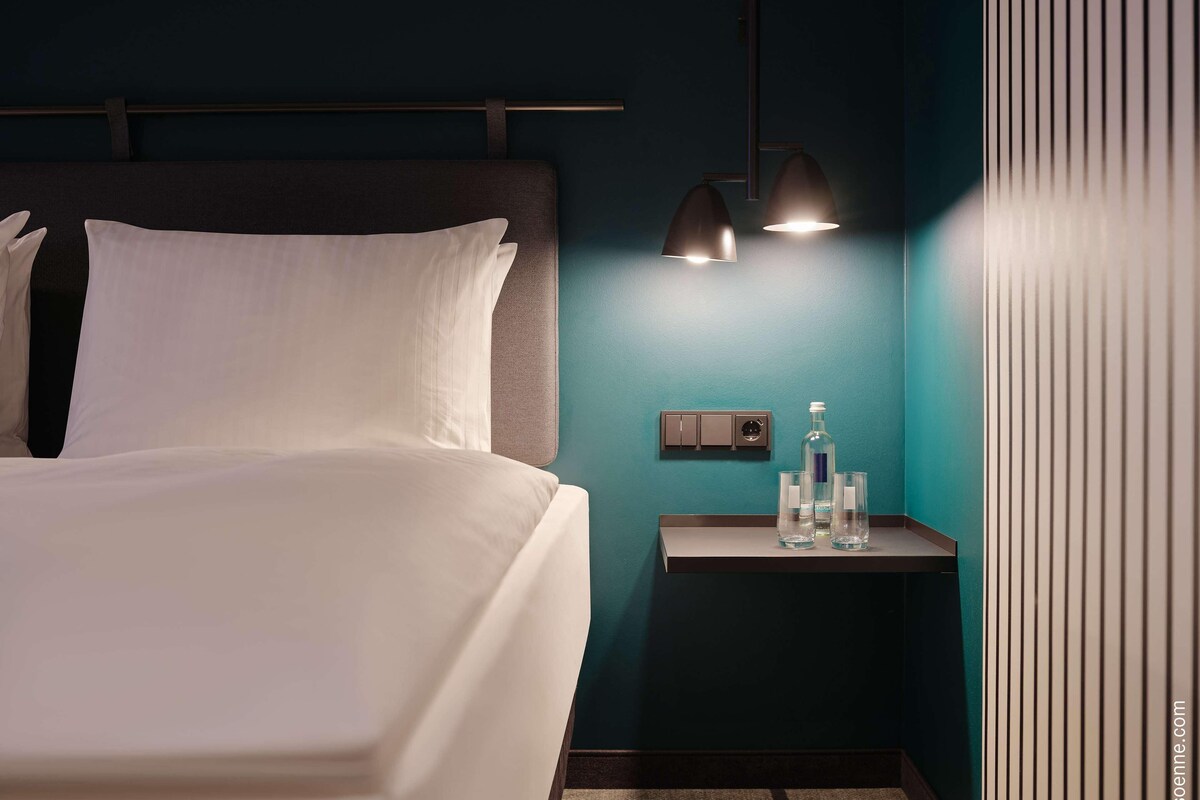 A modern hotel room with a neatly made bed, a nightstand with a bottle and glasses, and a wall-mounted power outlet.
