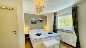 A bedroom with a bed, bedside lamps, a window with curtains, a radiator, and a ceiling light fixture. - Vacation apartment with a view (Sankt Englmar)