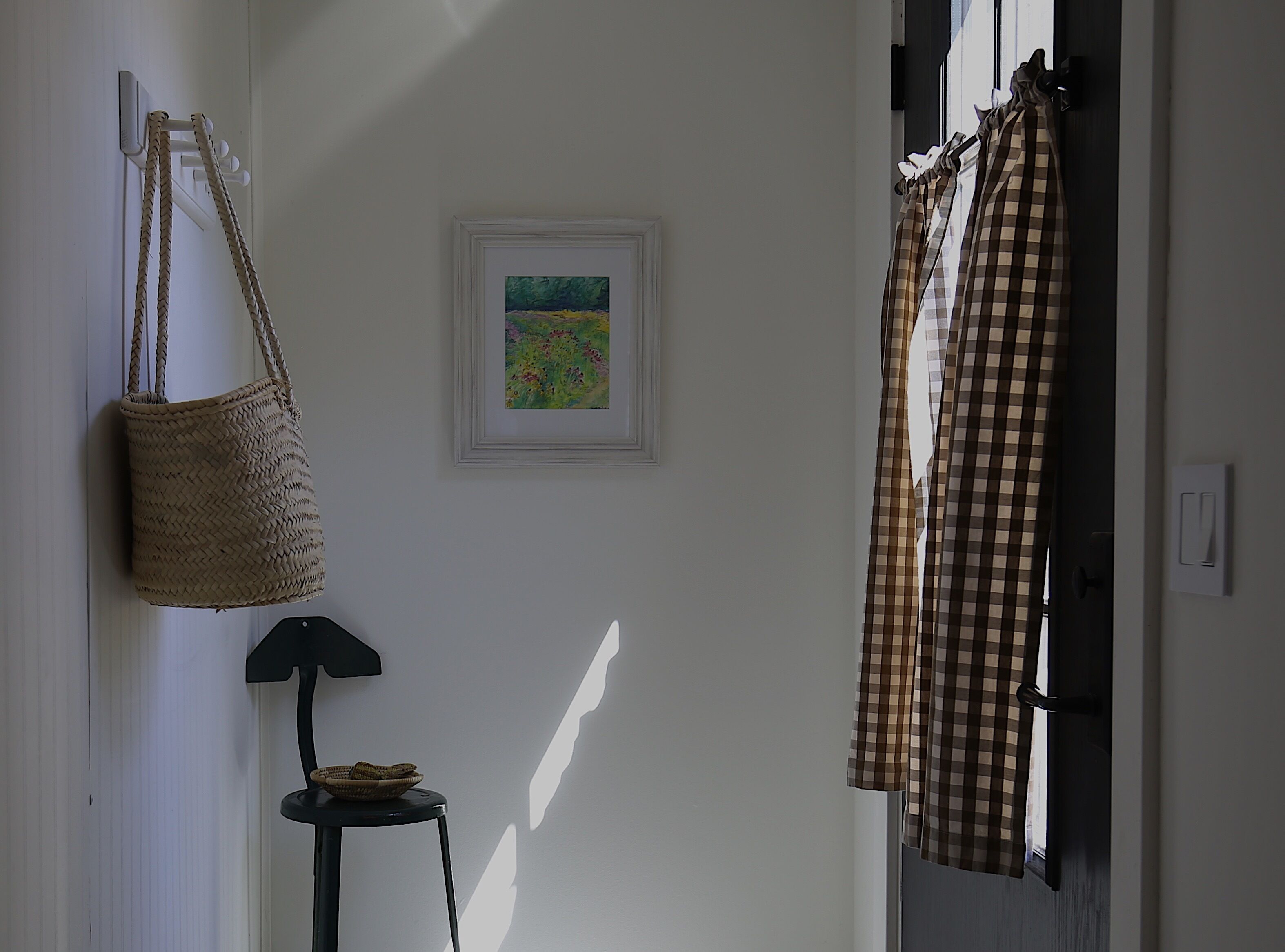 A hallway with a woven basket hanging on the wall, a framed painting, and a checkered curtain partially drawn.