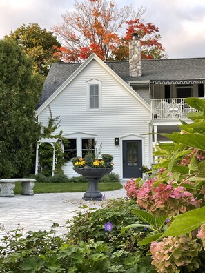 A white house with a stone chimney, a balcony with a railing, and a garden with a large planter and hydrangeas. - Boutique Studio in the Heart of Charlevoix (Charlevoix)