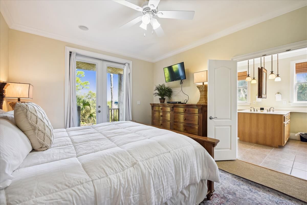 A bedroom with a large bed, a ceiling fan, a TV, a wooden dresser, and a bathroom with a sink and mirror.