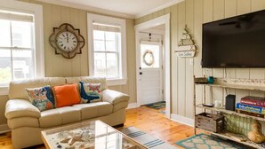 A living room with a beige sofa, a glass coffee table, a wall clock, a television, and a shelf with books and decorative items. - Located close to wedding venues, university, shops and eateries and Newport.   (Bristol)